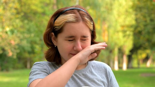 A teenage girl with dyed hair close-up sneezes and blows her nose in a paper napkin against the background of green nature