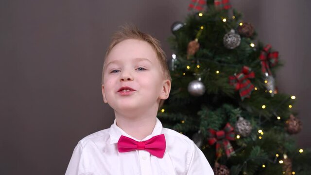 Positive little boy in white shirt with red bow tie talks standing near stylish decorated Christmas tree and grey wall in room