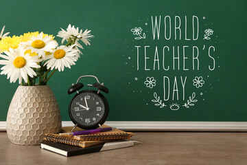 Vase with flowers, alarm clock and stationery on table near blackboard with text WORLD TEACHER'S DAY