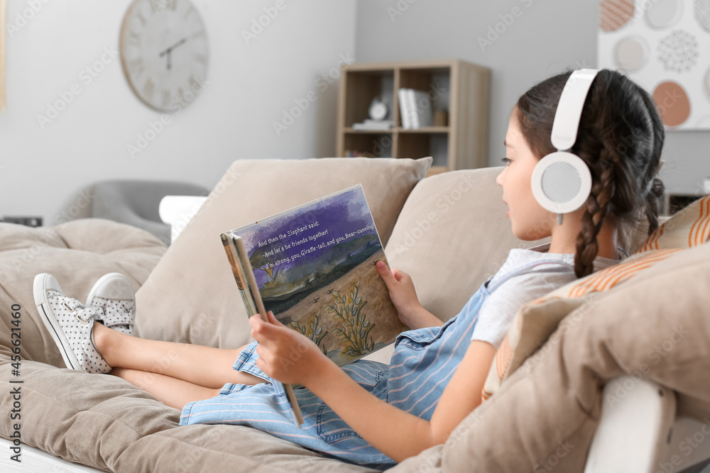 Wall mural Little girl with headphones reading book while sitting on sofa