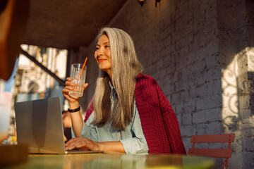 Dreamful senior Asian lady with long hair holds glass sitting at small table with open laptop on outdoors cafe terrace at sunset