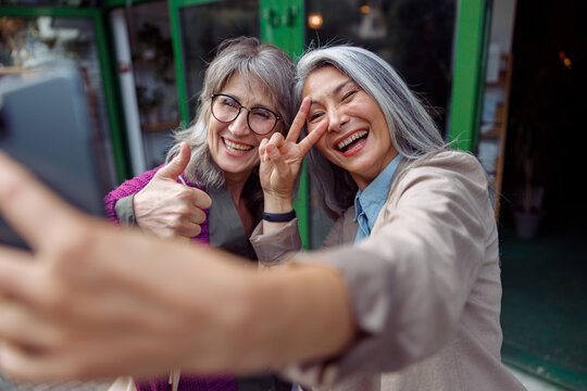 Attractive smiling mature Asian woman with friend take selfie gesturing with mobile phone on modern city street. Long-time friendship relationship