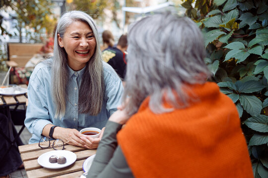 Laughing mature Asian woman with grey haired friend sit meeting at small table on outdoors cafe terrace on autumn day