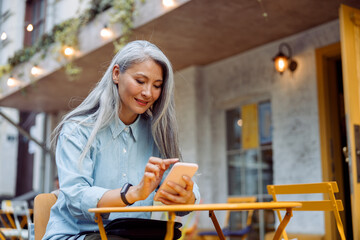 Positive grey haired senior Asian woman surfs internet with mobile phone sitting at yellow table on outdoors cafe terrace on autumn day