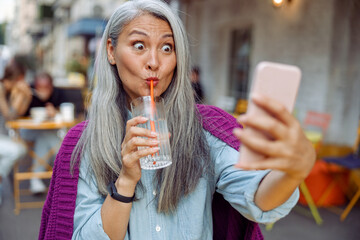 Funny mature Asian woman with long hair drinks water through straw taking selfie with modern...