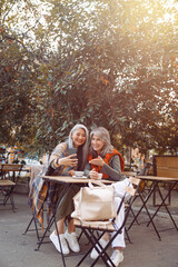 Happy grey haired female friends talk at videochat via cellphone sitting at small table in street cafe on nice autumn day