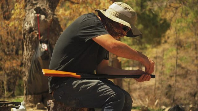 A man sits and reloads his bb gun while hunting for sport in a forest