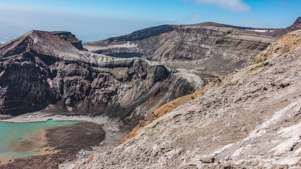 The caldera of an active volcano. The structure of the steep slopes of the crater is visible. At the bottom is a turquoise acid lake with melted snow on the banks. Blue sky. Kamchatka. Uzon