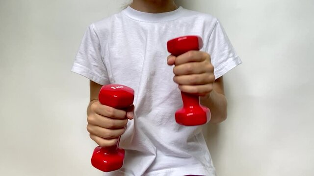 A girl in a white T-shirt slowly moves two red dumbbells weighing 1 kg 