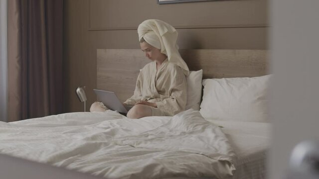 Stab shot of young woman wearing bathrobe and towel on head working on laptop computer sitting in bed in modern apartment