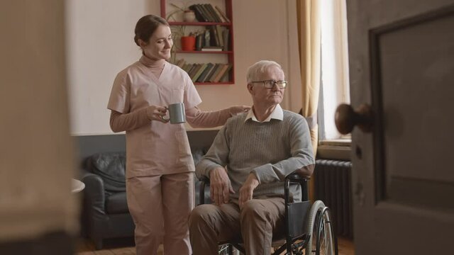 Slowmo shot of young supportive nurse bringing hot tea to old man in wheelchair, taking care of him at home for the elderly