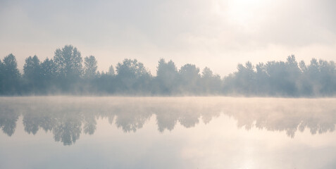 Beautiful foggy morning. Lake coast trees symmetrically reflect in the river water. Fog over autumn lake water.