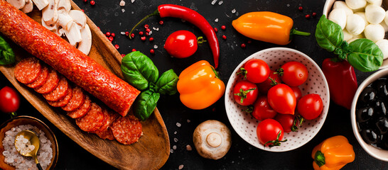Web banner with pizza ingredients on the dark gray background. Pepperoni sausage, mozzarella cheese, tomatoes, olives, mushrooms and flour are different products for making pizza and pasta