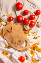 On the tablecloth are scattered colored pasta, tomatoes and garlic around an olive tree cutting board. 