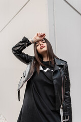 Fashion hipster girl in fashionable leather rock clothes with jacket and sweatshirt poses near the gray wall in the city and looks at the camera