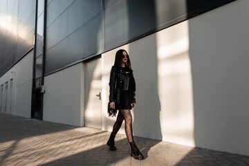 Naklejka premium Fashion girl model with sunglasses in trendy black clothes with leather jacket and handbag in sexy tights with shoes walks on the street near a modern building at sun light. Urban female style