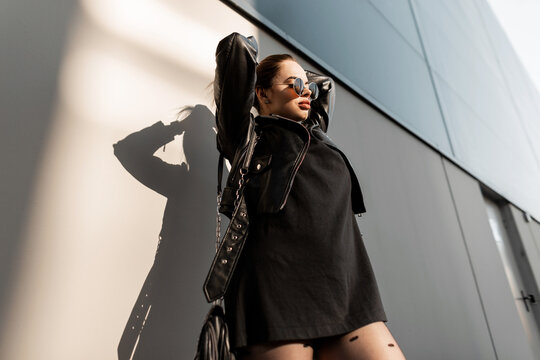 Fashionable hipster woman with vintage round sunglasses in stylish black clothes with leather jacket and dress with handbag stands near wall in city in sunlight