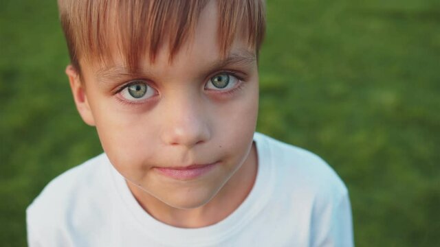 A little boy is staring intently into the camera with a slight squint. Close up of his face. He has kind, gray eyes. A bang falls on his forehead. Child in a white T-shirt