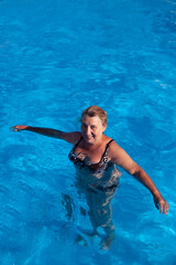 Woman is standing in water. Blonde with wet hair in beautiful swimsuit stands in pool and looks at camera. 