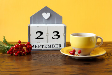 Calendar for September 23 : the name of the month in English, cubes with the number 23, a yellow cup of tea on a saucer, a branch of viburnum on a wooden table, a yellow background, side view
