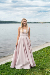 Fototapeta na wymiar Young woman in long pink evening dress standing alone near lake at city park