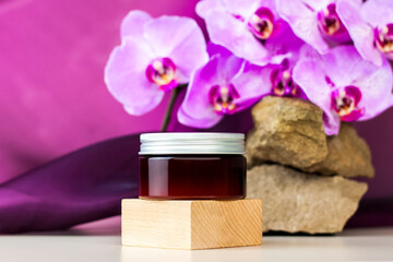 A brown jar of cream stands on a wooden pedestal against a stone background with beautiful orchids next to it. Stylish appearance of the product, layout, personality.