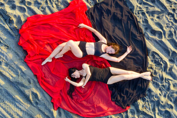 Two lovely girls in black bathing suits lie on red and black fabric on sandy beach. View from above