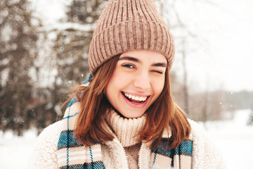 Young beautiful smiling hipster female in trendy warm clothes and scarf.Carefree woman posing in the street in park. Positive pure model having fun in snow. Enjoying winter moments. Winking