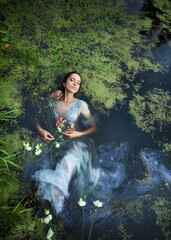 Art beautiful romantic woman lies in swamp in blue long dress with flowers. Portrait brunette in...