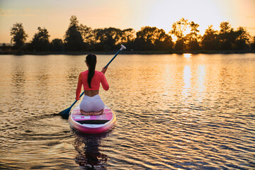 Back view of young woman in sportswear sitting on sup board with long paddle and enjoying beautiful...