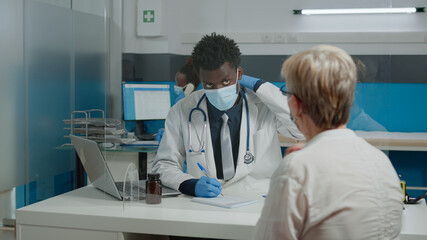 Obraz premium Medic with white coat sitting at desk while talking to elder woman in pain. Doctor doing healthcare checkup wearing face mask, gloves and stethoscope in medical cabinet at facility