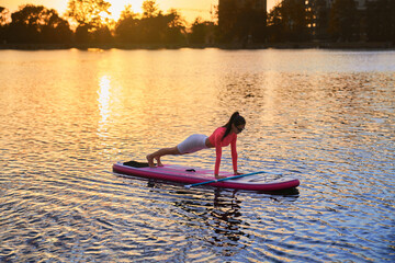 Strong young woman standing in plank position on sup board. Slender brunette training abs on city...