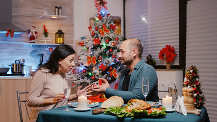 Man proposing with engagement ring to woman at festive dinner. Couple getting engaged while celebrating christmas eve with chicken meal and glasses of champagne. Cheerful people in love