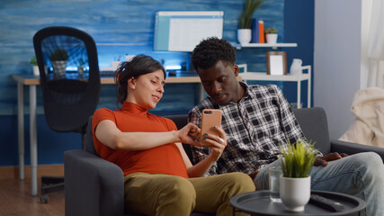 Married interracial couple relaxing on living room sofa at home. Pregnant caucasian woman using...