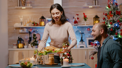 Festive couple preparing for christmas eve dinner at home. Woman bringing plate with seasonal food at table while man sitting to eat. People celebrating holiday with meal and champagne