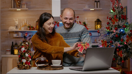 Festive couple giving presents on video call conference for christmas eve festivity. People talking to family on laptop and exchanging gifts on camera for remote seasonal celebration.