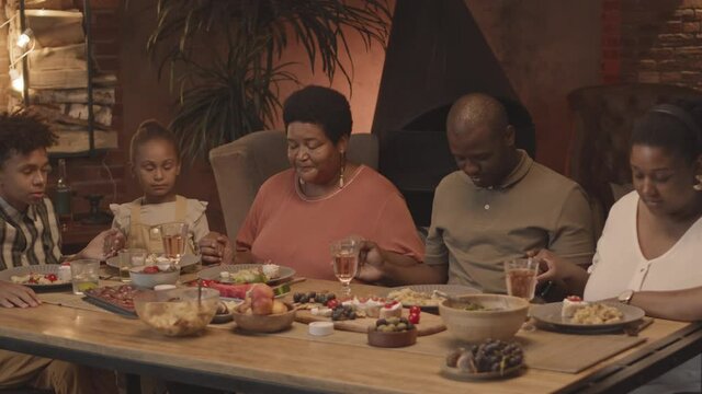 Medium close-up of African-American family sitting at table in dining room at home, holding hands and praying before meal