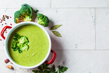 Broccoli soup puree in red saucepan on gray tile background with ingredients. Cooking healthy food...