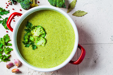 Broccoli soup puree in red saucepan on gray tile background with ingredients. Cooking healthy food concept.