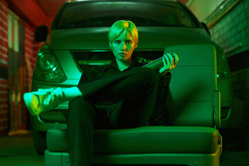 a girl with short hair sits in the garage near the car and holds a bat in her hand. Modern photo in neon green light. High quality photo