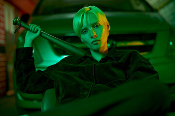 fashionable shooting in neon green light girl in a tracksuit sits near the car and holds a bat behind her back. High quality photo