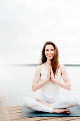 Fototapeta na wymiar Portrait of beautiful Caucasian woman in white clothes practising yoga on a lake shore