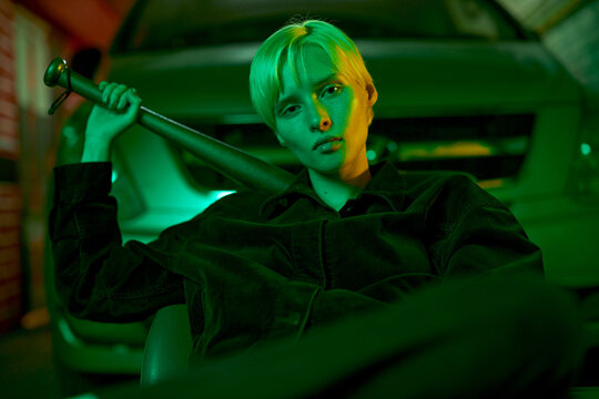 fashionable shooting in neon green light girl in a tracksuit sits near the car and holds a bat behind her back. High quality photo