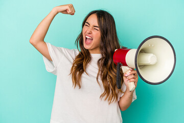 Young caucasian woman holding a megaphone isolated on blue background raising fist after a victory,...