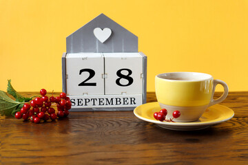 Calendar for September 28 : the name of the month in English, cubes with the number 28, a yellow cup of tea on a saucer, a branch of viburnum on a wooden table, a yellow background, side view