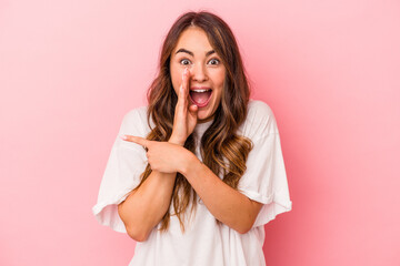 Young caucasian woman isolated on pink background saying a gossip, pointing to side reporting something.