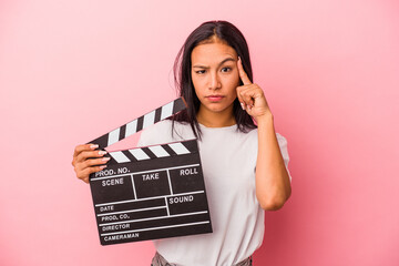 Young latin woman holding clapperboard isolated on pink background  pointing temple with finger,...
