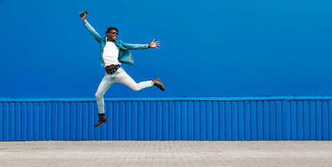 african guy jumping with a phone in his hands, in the city against the background of a blue building
