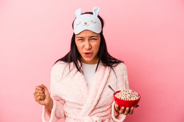 Young latin woman wearing pajama holding a bowl of cereals isolated on pink background  screaming very angry and aggressive.
