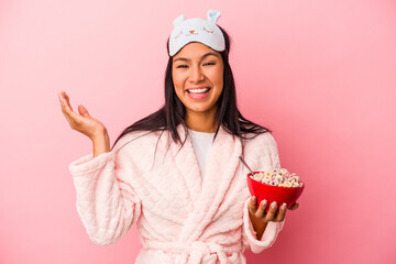 Young latin woman wearing pajama holding a bowl of cereals isolated on pink background  receiving a pleasant surprise, excited and raising hands.